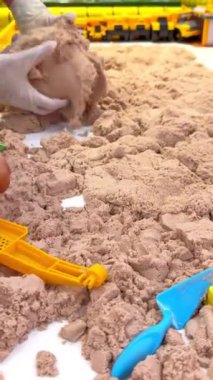 dikey video, çocukların dokunsal ve kinestetik gelişimi için kinetik kumla boks yapan çocuklar için çocuk parkı, Montessori, kauçuk eldivenli kadın elleri kum karıştırıyor, çocuk dereleri oynuyor