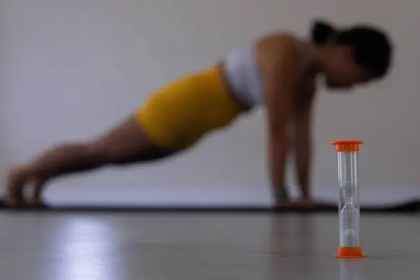 Turuncu kum saati yakın plan, arka plan, spor sınıfı, yoga, pilates ve fitness stüdyosunda sarı ve beyaz spor kıyafetleri içinde bulanık Koreli kadın. 