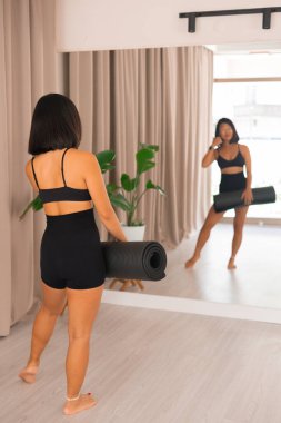 Çekici Asyalı spor eğitmeni siyah minderle ayakta duruyor ve aynaya bakıyor spor salonunda ve yoga stüdyosunda..