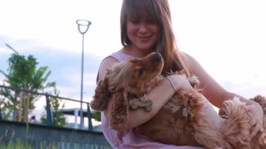 Genç ve güzel bir kadın cocker köpeğine sarılıyor ve öpüyor, sevimli evcil hayvan kameradan izliyor, yaz günbatımı halk parkında, yavaş çekim.