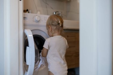 Ev işleri yapan iki yaşında bir çocuk. Çamaşır makinesi yükleniyor. Küçük kız evde çamaşır yıkamaya yardım ediyor.
