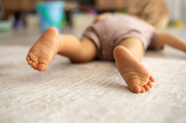 Close-up of baby feet, toddler lying on the floor at home, blurred toys and home interrior on background