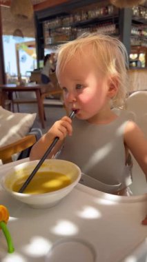 Küçük sevimli sarışın kız kafede pipetle püre çorba içiyor. Restauranttaki yazlık terasta güneş ışınları masaya düşüyor ve kızlar yüzüne bakıyor. Çocuklar için sağlıklı beslenme ve