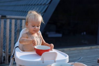 Sevimli sarışın kız terasta beyaz bir bebek sandalyesinde oturuyor ahşap bir evin terasında ve kırmızı bir tabaktan yulaf lapası yiyor, çocuk gülümsüyor ve iştahla yemeğini yiyor.