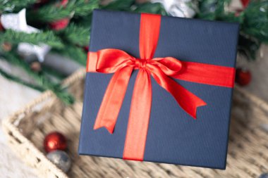 Kırmızı kurdeleli siyah hediye kutusu, Yeni Yıl 2025 ve Noel hediyesi, köknar ağacı ve süsleme topları arka planda, tatil konsepti