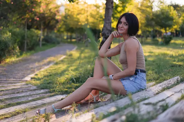 stock image Portrait of a young pretty woman, sitting on a wooden stairs in green public park during evening summer sunset. Smiling, posing and wathing in camera, beautifful rays of sun and shadows on background