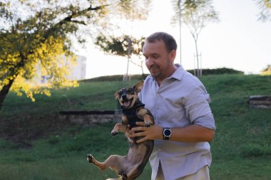 Gülümseyen orta yaşlı bir adam halka açık bir parkta küçük bir köpekle yürüyor ve oynuyor, barınaktan evlat edinilmiş çok cins bir fare teriyeri, birlikte mutlu zaman, aktiviteler ve bir evcil hayvanla açık hava oyunları,