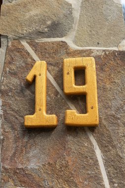 Taş bir duvarda on dokuz numaralı sarıya yakın çekim. Parlak, ahşap, turuncu figür 19. Kır otelinde bir ev veya oda numarası. Doğal malzemeler ve çevre dostu stil. Yüksek kalite fotoğraf