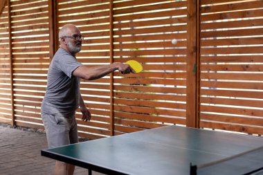 Gri saçlı, gülümseyen orta yaşlı adam masa tenisi oynuyor. Elinde sarı bir raket tutuyor ve beyaz bir top atıyor. Ahşap bahçe evi ve arka planda tahta çit. Kır evinde dinlenin ve dinlenin.