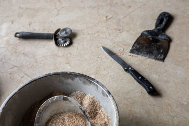 Çeşitli fırın ve makarna üretim araçları ve malzemeleri, bir kase un, ölçüm kabı ve kazıyıcı sergilenen, sade bir mutfak tezgahına yerleştirilmiştir.