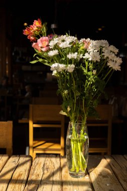 Beyaz papatyalar ve pembe alstroemeria da dahil olmak üzere canlı bir çiçek düzeni, kırsal bir ahşap masadaki şeffaf cam bir vazoda zarif bir şekilde duruyor. Sıcak güneş ışığı süzülüyor, yumuşak dökülüyor.