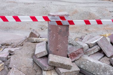 Bir inşaat sahasında dağınık tuğla ve taşlarla çevrili dikey bir beton direk bulunuyor. Kırmızı ve beyaz renklerde uyarı bandı direğin etrafına sıkıca bağlanmış. Bu da yasak bölge anlamına geliyor.