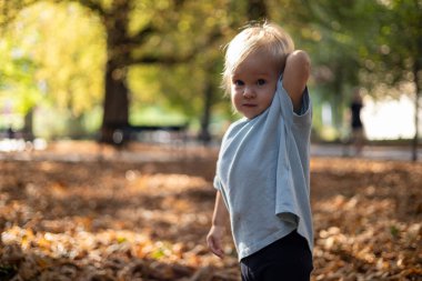 Neşeli bir çocuk, canlı sonbahar yapraklarıyla çevrili bir parkta güneşli bir günün tadını çıkarıyor. Küçük kız altın yapraklardan bir halının ortasında duruyor, merak ve neşe dolu bir ifadeyle. Yumuşak