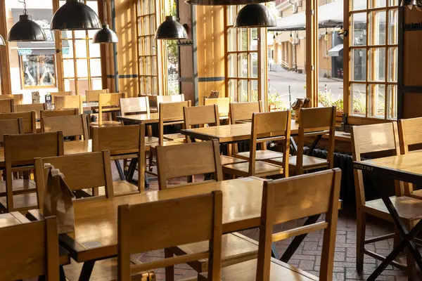 Konforlu bir kafe içinde geniş pencerelerden gelen sıcak, doğal ışıkla yıkanmış basit ahşap sandalyeler ve masalar bulunur. Müşterilerin yokluğu barışçıl bir ortam yaratırken,