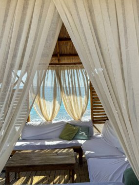 Sakin bir plaj kabininde rüzgarda sallanan beyaz perdeler, yumuşak yastıklarla süslenmiş geniş bir oturma alanını çerçeveler. Batan güneş okyanus üzerinde sıcak bir parıltı yayar ya da