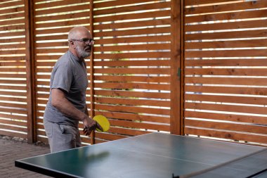 Gri saçlı, gülümseyen orta yaşlı adam masa tenisi oynuyor. Elinde sarı bir raket tutuyor ve beyaz bir top atıyor. Ahşap bahçe evi ve arka planda tahta çit. Kır evinde dinlenin ve dinlenin.