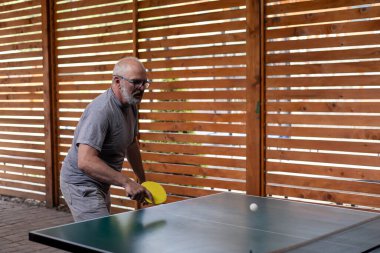 Gri saçlı, gülümseyen orta yaşlı adam masa tenisi oynuyor. Elinde sarı bir raket tutuyor ve beyaz bir top atıyor. Ahşap bahçe evi ve arka planda tahta çit. Kır evinde dinlenin ve dinlenin.