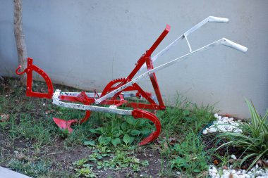 Kırmızı ve beyaza boyanmış eski bir bahçe sabanı, küçük kayalar ve bitkilerle çevrili yemyeşil bir bahçede boşta duruyor. Klasik bir tasarım sergileyen bu rustik ekipman, geleneksel