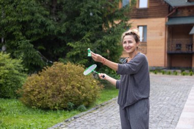 Güzel gülümseyen orta yaşlı bir kadın badminton oynar. Elinde yeşil bir raket ve küçük bir horoz tutuyor. Ahşap bahçe evi ve arka planda tuğla ev. Kır evinde ya da otelde dinlenin.