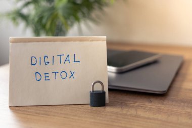 DiGITAL DETOX etiketli el yazısıyla yazılmış bir not rustik ahşap bir masanın üzerinde duruyor, fişi çekme ve ekranlara ara verme temasını vurguluyor. Notun yanında, küçük bir asma kilit akıllı bir telefonu güvenceye alıyor.