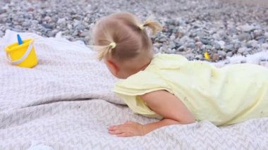 Genç sarışın bir çocuk kumsalda bir battaniyeye uzanıyor, yanında sarı bir kova otururken renkli çakıl taşlarına bakıyor. Küçük kız çığlık atıyor ve gülüyor. Bu mükemmel günde güneş parlıyor.