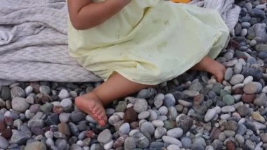 Sahilde taş ve deniz kabuklarıyla oynayan bebek ayaklarının yakın çekimi. Sarı elbiseli genç bir kız, çakıl taşı kaplı bir plajda battaniyenin üzerinde oturuyor, yeni yürümeye başlayan çocuk mutlu bir şekilde oyun oynuyor ve dokularının tadını çıkarıyor.