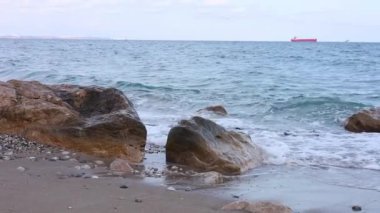 Güneş, yumuşak dalgaların kayalara çarpmasıyla güzel bir kumsalda yükseliyor ve suyun yanında huzurlu bir atmosfer yaratıyor. Uzaklarda bir kargo gemisi sürükleniyor..