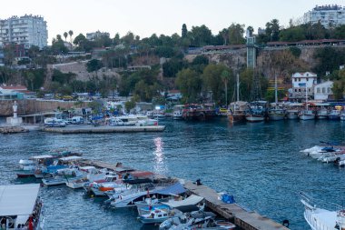 Antalya, Türkiye - 28.09.2024: Bereketli yeşil tepeler ve modern binalar tarafından çerçevelenmiş hareketli bir marinanın sakin suyunda renkli tekneler yavaşça sallanıyor. Batan güneş sahnenin üzerine ılık bir parıltı saçar.