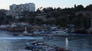 Antalya, Türkiye - 28.09.2024: Bereketli yeşil tepeler ve modern binalar tarafından çerçevelenmiş hareketli bir marinanın sakin suyunda renkli tekneler yavaşça sallanıyor. Batan güneş sahnenin üzerine ılık bir parıltı saçar.