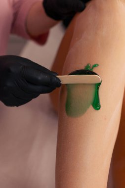 Siyah eldiven giyen bir güzellik uzmanı kuaför salonundaki bir müşterinin bacağına yeşil mum sürer. Bacak, beyaz bir tedavi masasının üzerine yerleştirilmiş. Saç kesme tekniğini vurguluyor. Yumuşak ışıklandırma