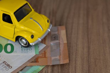 Küçük, parlak, sarı bir oyuncak araba, ahşap bir masa yüzeyine karşı kurulmuş bir kaç Euro banknotunun üzerine yerleştirilmiştir. Düzenleme, finans, para ve ulaşım konularında eğlenceli bir yaklaşım öneriyor..