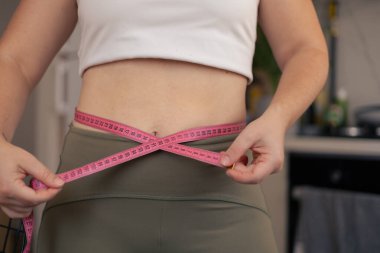 Üniformalı bir kadın mutfakta durmuş, kemerini pembe bir mezurayla ölçüyor, fitness hedeflerine ve ilerlemesine odaklanıyor. Kilo kaybı, diyet ve vücut pozitif konsept