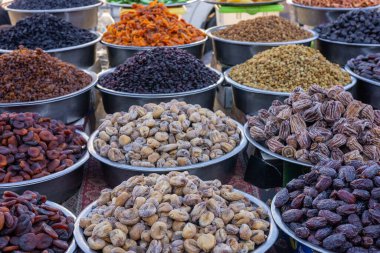 Kalabalık bir pazarda bir dizi kurutulmuş meyve ve fındık sayısız kaseyi dolduruyor. Sahne canlı, öğleden sonra güneşinin altında birçok kumaş ve renk sergileniyor, müşterileri cezbediyor..