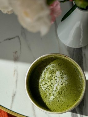 Enjoying a vibrant cup of lavender matcha latte or tea topped with frothy cream, paired with delicate pink flowers in a simple vase, all set on a stylish marble table. Modern cafe. Coffee alternative clipart