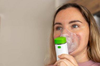 Genç bir kadın solunum tedavisi için nebulizör kullanıyor. Cihazı yüzüne yakın tutuyor, sağlık ve sağlığın önemini vurguluyor. Mevsimsel soğuk algınlığı, öksürük, boğaz ağrısı, burun akışı.
