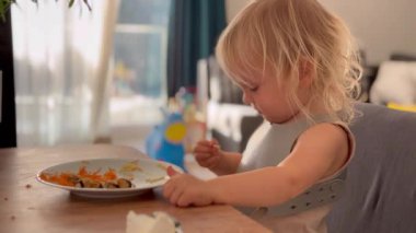 Küçük sarışın bir kız yemek masasında oturuyor, tabağında spagetti ile yemeğinin tadına bakıyor. Meşgul ve meraklı görünüyor, evde yemeğinin tadını çıkarırken lezzetleri keşfediyor..