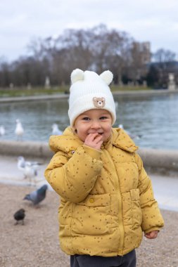 Parlak sarı bir ceket ve rahat beyaz bir şapka giyen bir çocuk Paris 'te bir parktaki çeşmenin yanında duruyor. Kuşlar suyun etrafında uçuyor ve toplanıyor, canlı bir atmosfer yaratıyor..
