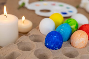 A person carefully paints a blue color on an Easter egg using a brush while candles glow softly nearby. The festive atmosphere is enhanced by spring flowers and a palette of vibrant colors. clipart