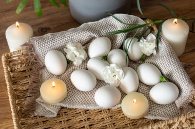 Dekoratif beyaz yumurtalar, narin çiçekler ve yumuşak mumlarla dokunmuş bir tepside sanatsal olarak düzenlenir. Baharda Paskalya bayramını kutlarken ortam huzurlu bir atmosfer yayıyor..