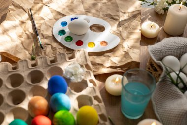 Paskalya için bahar çiçekleri ve mumlarla süslenmiş bir masada renkli yumurtalar süsleniyor. Boyalı ve fırçalı bir palet yakında, bayram için şenlikli bir ortam yaratıyor. Güneşli bir gün