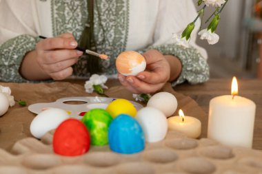 Bir insan, mum ve çiçeklerle çevriliyken Paskalya yumurtalarına renkli süsler çizer. Neşeli atmosfer bahar ruhunu ve bayram kutlamasını yansıtıyor..