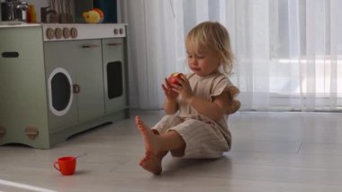 Kıvırcık sarı saçlı bir çocuk açık bir zeminde oturup taze kırmızı elma yer. Parlak alanda bir oyun mutfağı ve bölgeyi aydınlatan doğal ışık, oyuncu yaratıcılığı artırıyor..