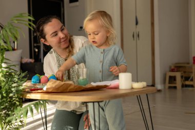 Bir anne ve çocuk evde neşeli bir Paskalya yumurtası süsleme seansına giriyorlar. Masada renkli yumurtalar sergileniyor, bayramın şenlikli ruhunu yansıtıyor..