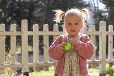 A young girl with pigtails delights in the Easter festivities, holding bright green egg while standing in a grassy area near a white picket fence, basking in the warm sunlight. Toddler girl in dress clipart