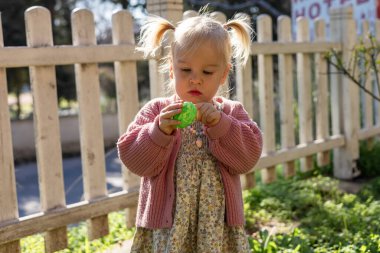 A young girl with pigtails delights in the Easter festivities, holding bright green egg while standing in a grassy area near a white picket fence, basking in the warm sunlight. Toddler girl in dress clipart