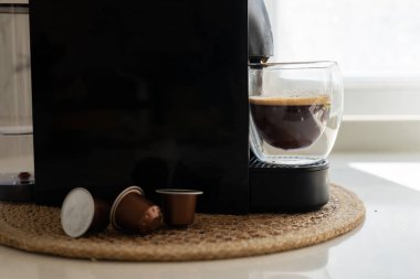 Çağdaş bir espresso makinesi şık bir cam fincanda zengin bir fincan kahve hazırlar. Kahve kapsülleri yakınlarda dokuma hasırının üzerinde oturur, kahve yapmanın basitliğini vurgularlar. Günlük rutin