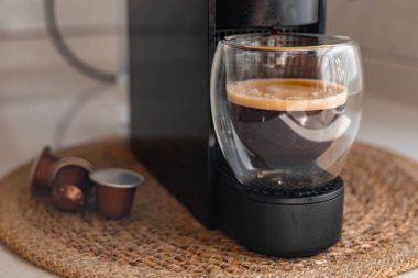 Çağdaş bir espresso makinesi şık bir cam fincanda zengin bir fincan kahve hazırlar. Kahve kapsülleri yakınlarda dokuma hasırının üzerinde oturur, kahve yapmanın basitliğini vurgularlar. Günlük rutin