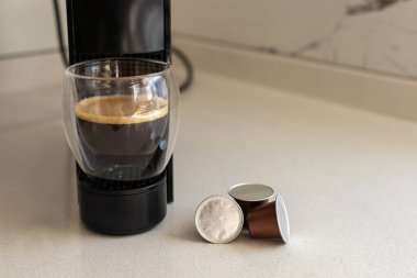 Gösterişli bir espresso makinesi çağdaş bir mutfakta bir fincan taze kahve hazırlar. Parlak sabah ışığı, kullanılmış kahve kapsüllerinin yanındaki zengin espresso bardağını vurgular.