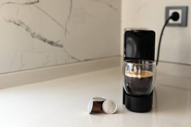 Gösterişli bir espresso makinesi çağdaş bir mutfakta bir fincan taze kahve hazırlar. Parlak sabah ışığı, kullanılmış kahve kapsüllerinin yanındaki zengin espresso bardağını vurgular.
