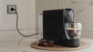 Şık, modern bir mutfakta, kapsül makinesi zengin bir espresso yapıp cam bardağa doldurur. Kurulumda, sabah evde ideal bir kahve anı gösteren kapsüller ve su deposu yer alıyor.
